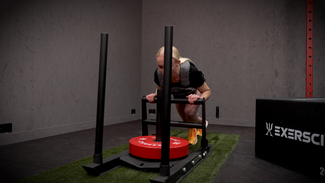 An image of a woman wearing an Exersci weighted vest pushing the Exersci® Weight Plate Sled Push to illustrate the blog post entitled 'Sled Training Benefits: Why And How To Complete It'