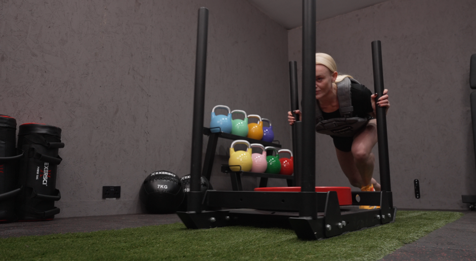 A woman completing a sled push with an Exersci weighted vest on in a home gym to illustrate the Exersci blog post entitled 'What Is The Essential Gym Equipment For A Home Gym?'