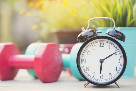 Alarm clock with dumbbells in the background to illustrate the Exersci blog post 'Is It Better to Exercise In The Morning Or Evening?'