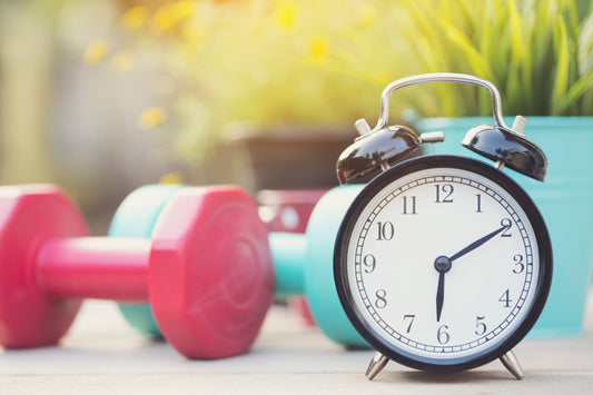 Alarm clock with dumbbells in the background to illustrate the Exersci blog post 'Is It Better to Exercise In The Morning Or Evening?'